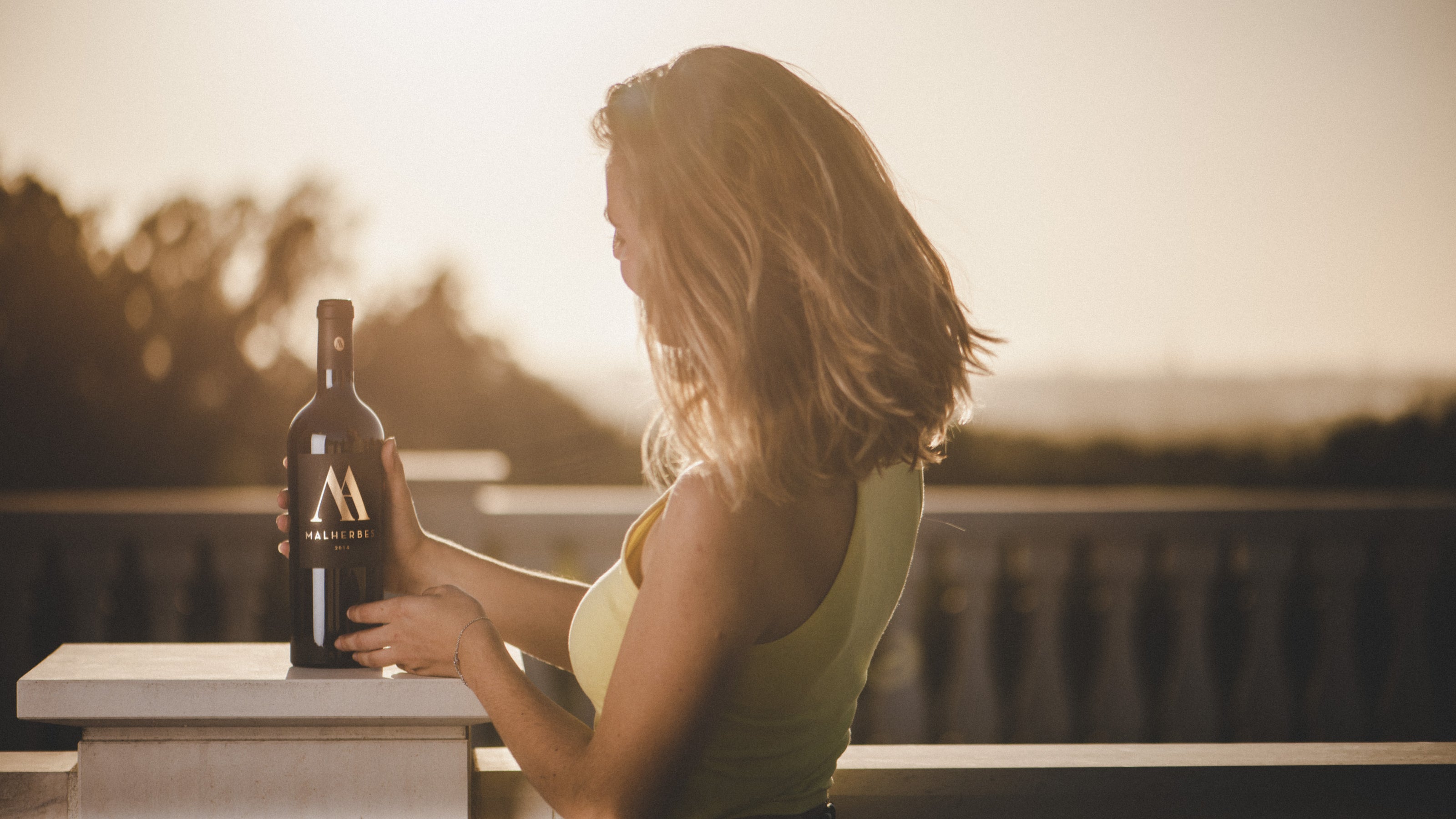 A woman standing on a terrace at the sunset holding a bottle of Malherbes Red Wine
